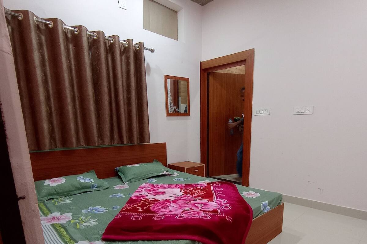 A bedroom with a wooden bed, green floral bedding, a red blanket, a small bedside table, a mirror, and a window with curtains.