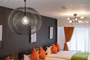Ein modernes Hotelzimmer mit einem großen Bett, orangefarbenen Kissen, einer hängenden, kugelförmigen Leuchte und einem Deckenventilator.
