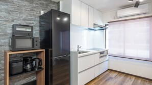 A modern kitchen with a built-in microwave, a black refrigerator, white cabinets, a wooden shelf with kitchen appliances, and a window with blinds. - Seeds Nipponbashi (Osaka)