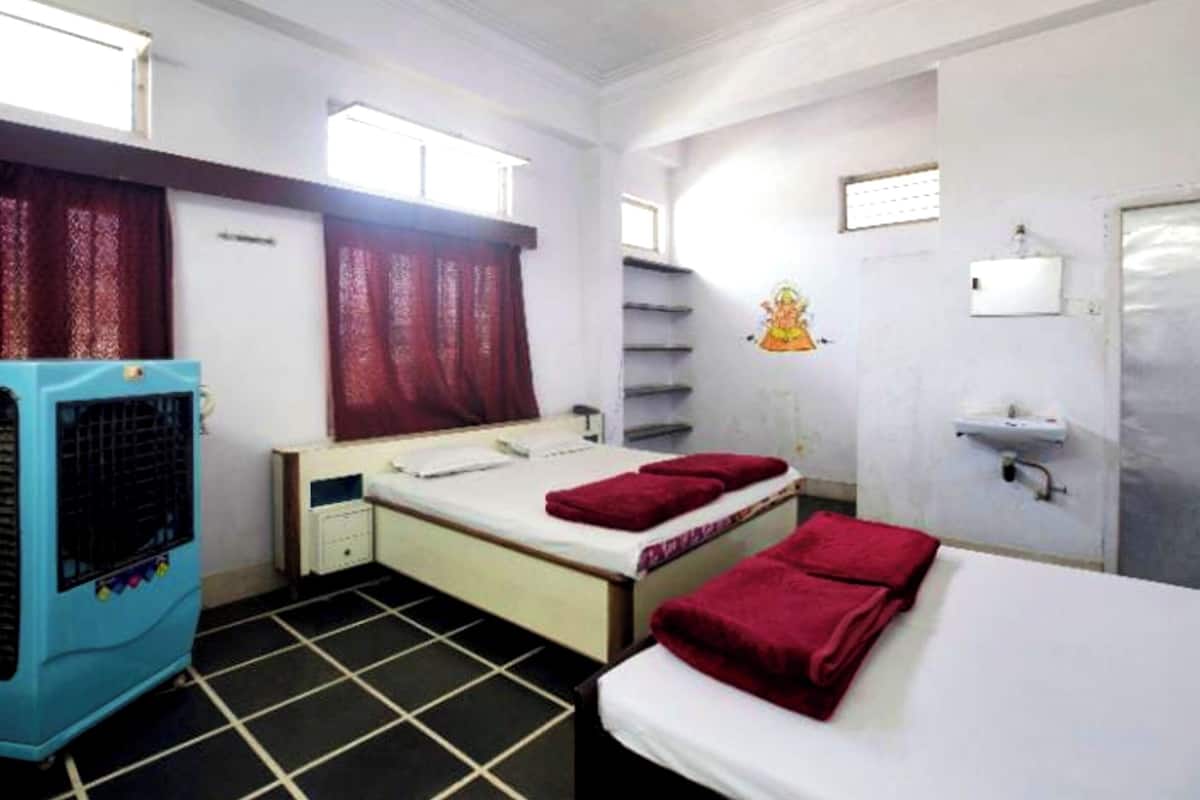 Une chambre d’hôtel avec deux lits, un radiateur, une fenêtre avec des rideaux, une étagère fixée au mur et un lavabo.
