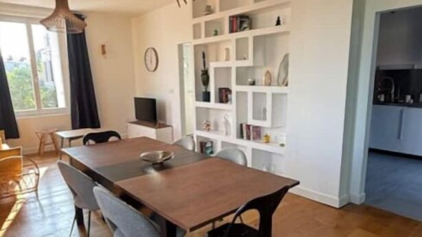 Un salon moderne avec une table à manger en bois, deux chaises, une bibliothèque intégrée et un coin cuisine avec des placards.