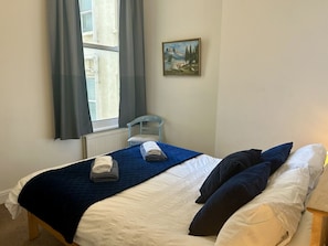 Un dormitorio con cama, una silla, una ventana con cortinas y un cuadro en la pared.