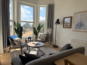 Een woonkamer met een bank, fauteuils, een ronde salontafel en een vloerlamp. Grote ramen met gordijnen, een radiator en een lijst met een afbeelding op de muur.