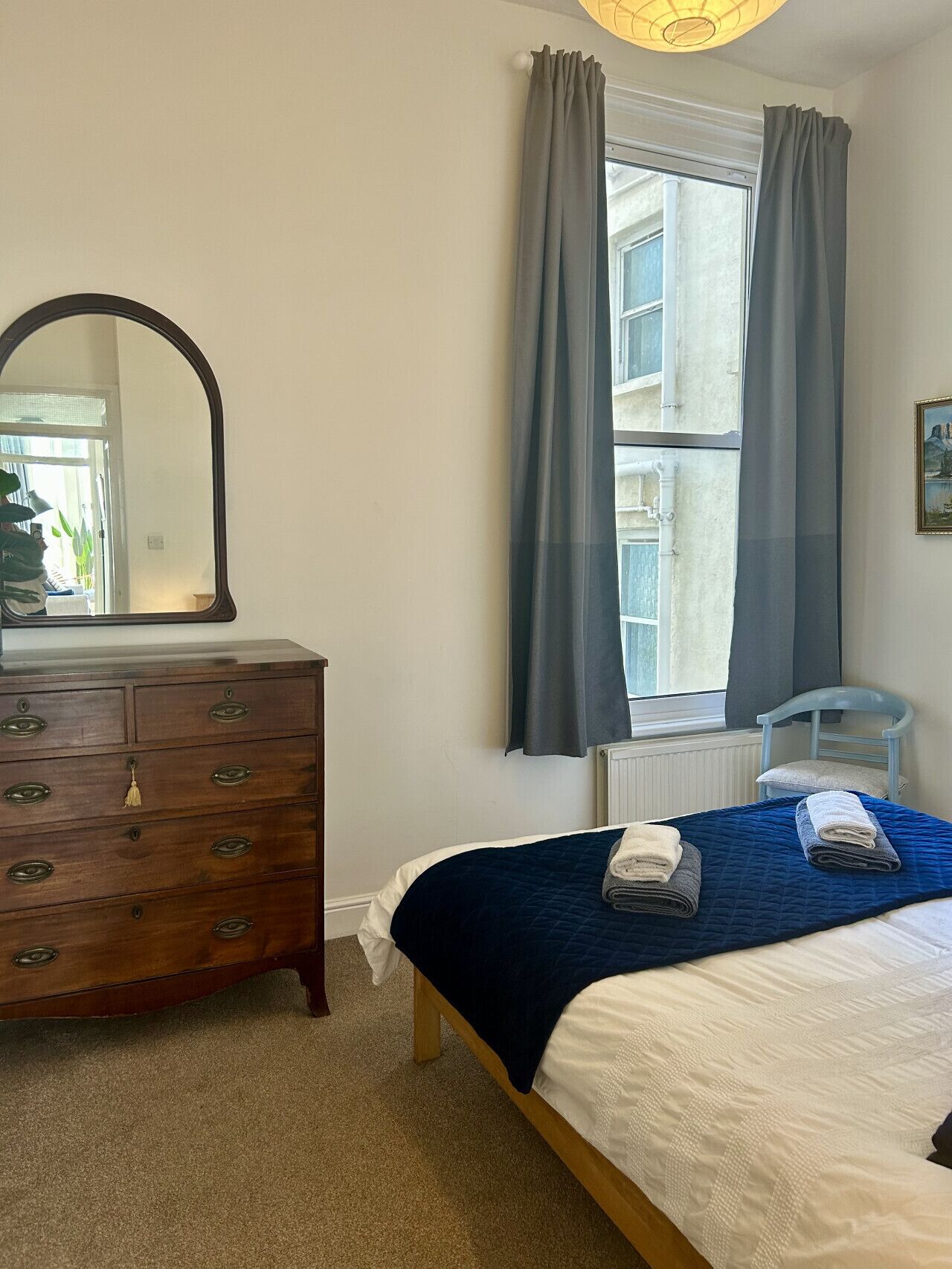 A bedroom with a bed, a dresser, a chair, a window with curtains, and a mirror.