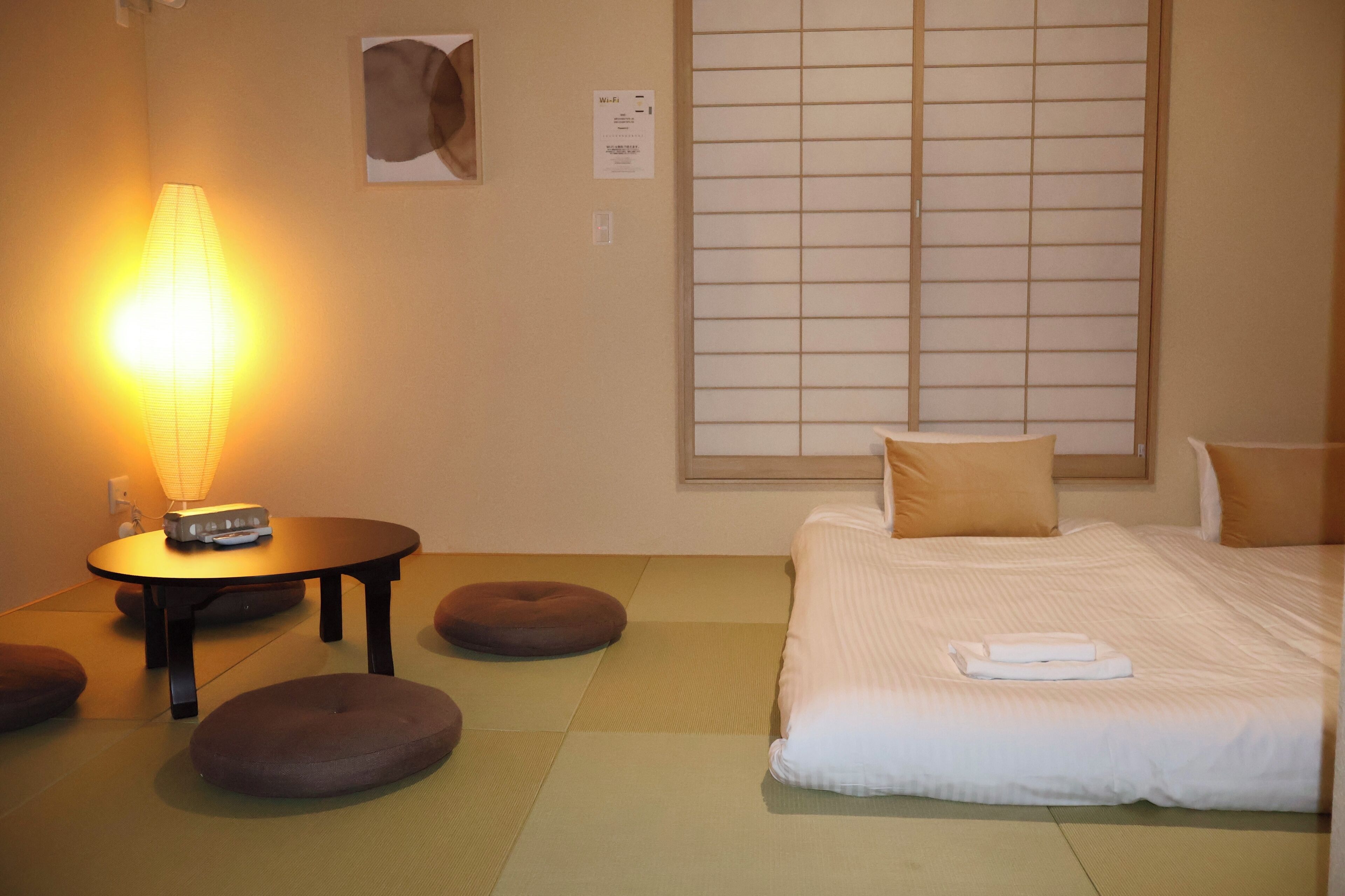 A hotel room with tatami flooring, a bed with white bedding, a bedside table with a lamp, and a window with a view of the outside.
