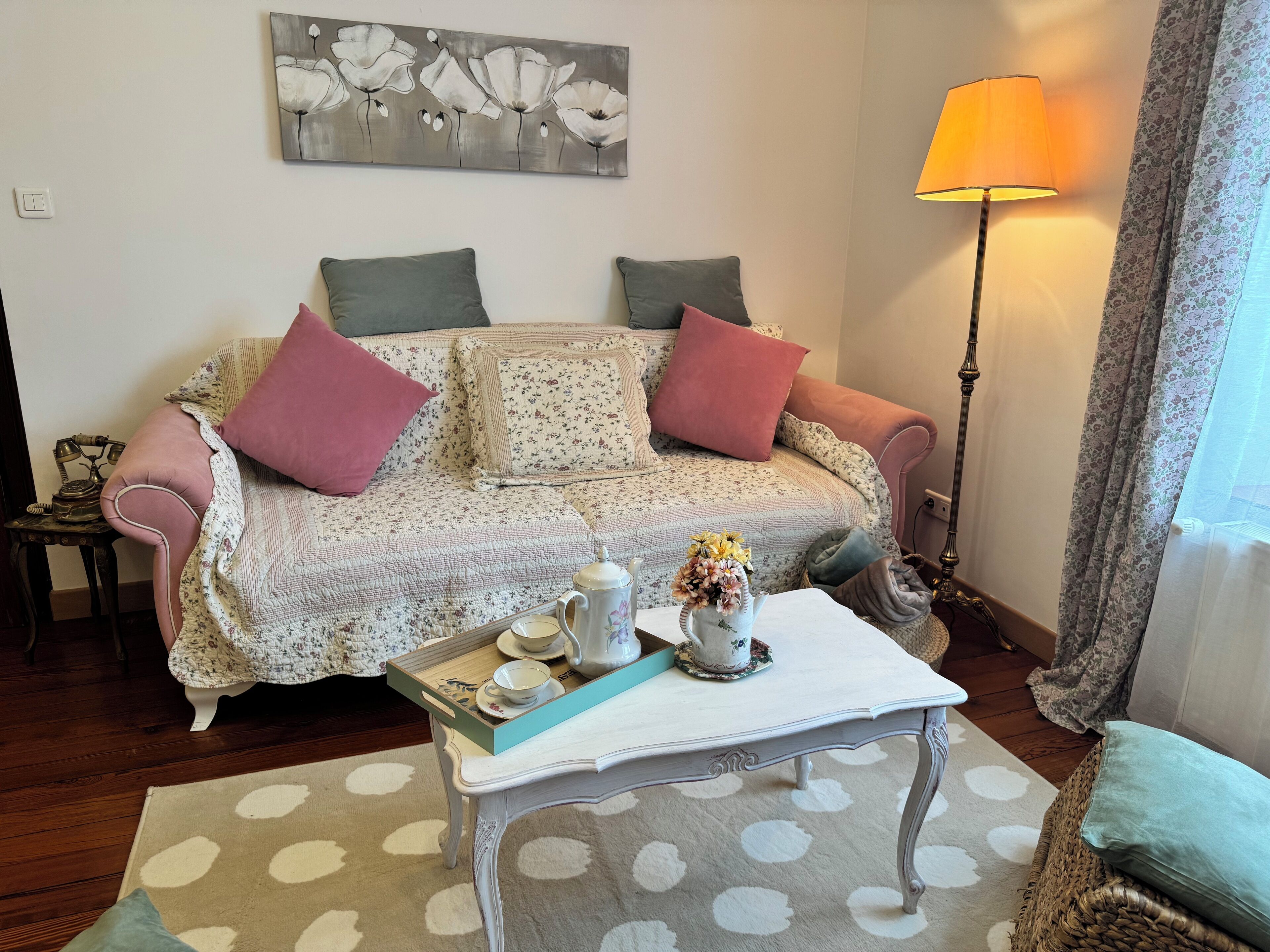 A cozy living room with a floral sofa, a small table with a teapot and cups, a lamp, and a painting on the wall.