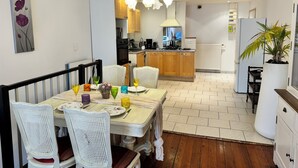 Un comedor con una mesa lista para cuatro personas, rodeada de sillas de mimbre blanco. Se ve una cocina al fondo, con armarios de madera y una estufa moderna.