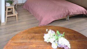 A bedroom with a wooden ceiling, a bed with pink bedding, a small stool with plants, and a wooden table with a vase of flowers. - Gîte "sous le préau (Cramans)
