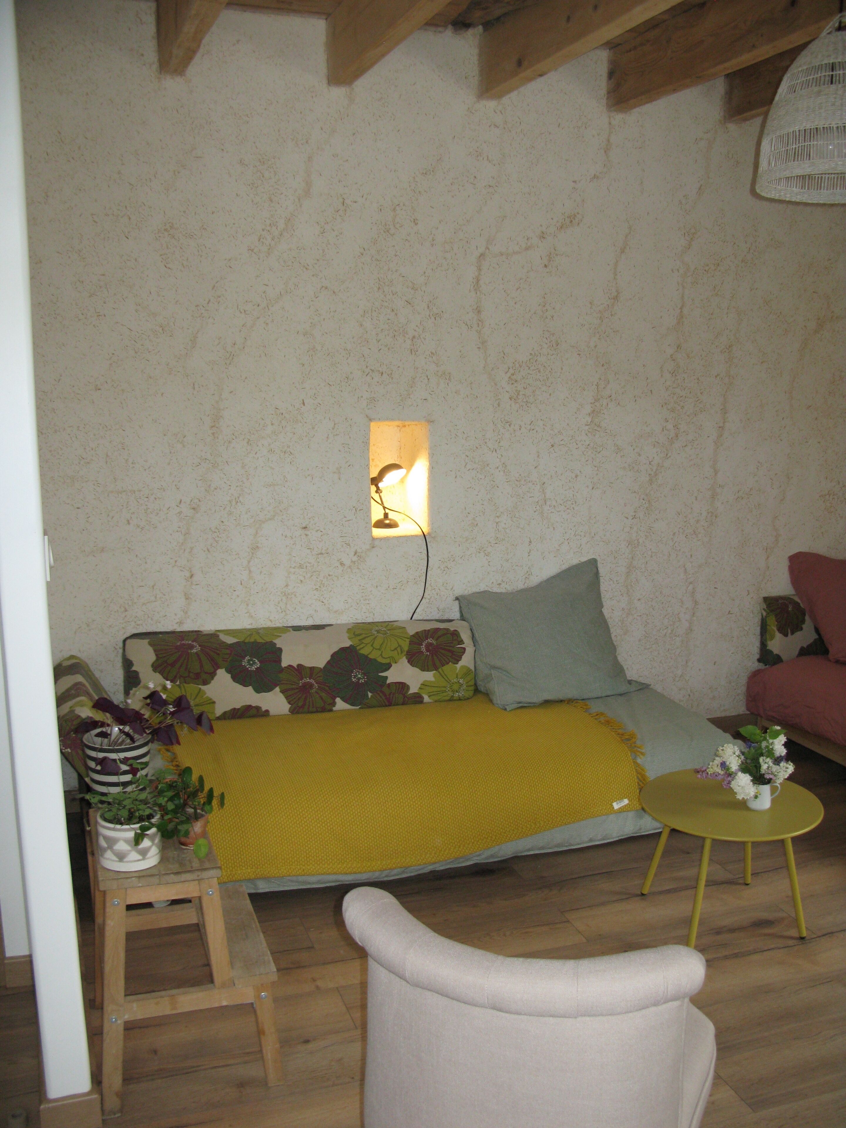 Un salon chaleureux avec un canapé, un petit tabouret en bois, un coussin jaune et une petite table ronde sur laquelle est posé un vase de fleurs.