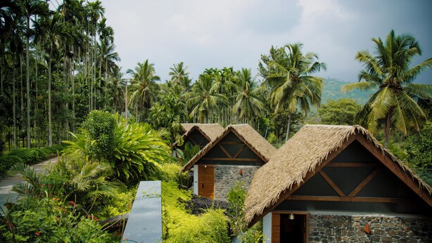 Two thatched-roof cottages surrounded by lush greenery and palm trees. - Maayaa riverside villa by KPM Tripenta (Mannarkkad)