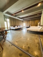 Una habitación de hotel amplia con una cama grande, una cama más pequeña y un sofá. Hay una mesa de madera y una silla. El piso es de concreto con un borde decorativo.