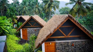 Two thatched-roof cottages with stone walls, surrounded by lush greenery and palm trees. - Maayaa riverside villa by KPM Tripenta (Mannarkkad)