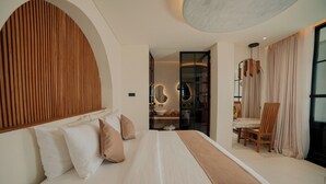 A modern hotel room with a large bed, a wooden headboard, a seating area with a table and chairs, and a bathroom with a mirror and sink. - OhJulia Hotel Komodo (Labuan Bajo)
