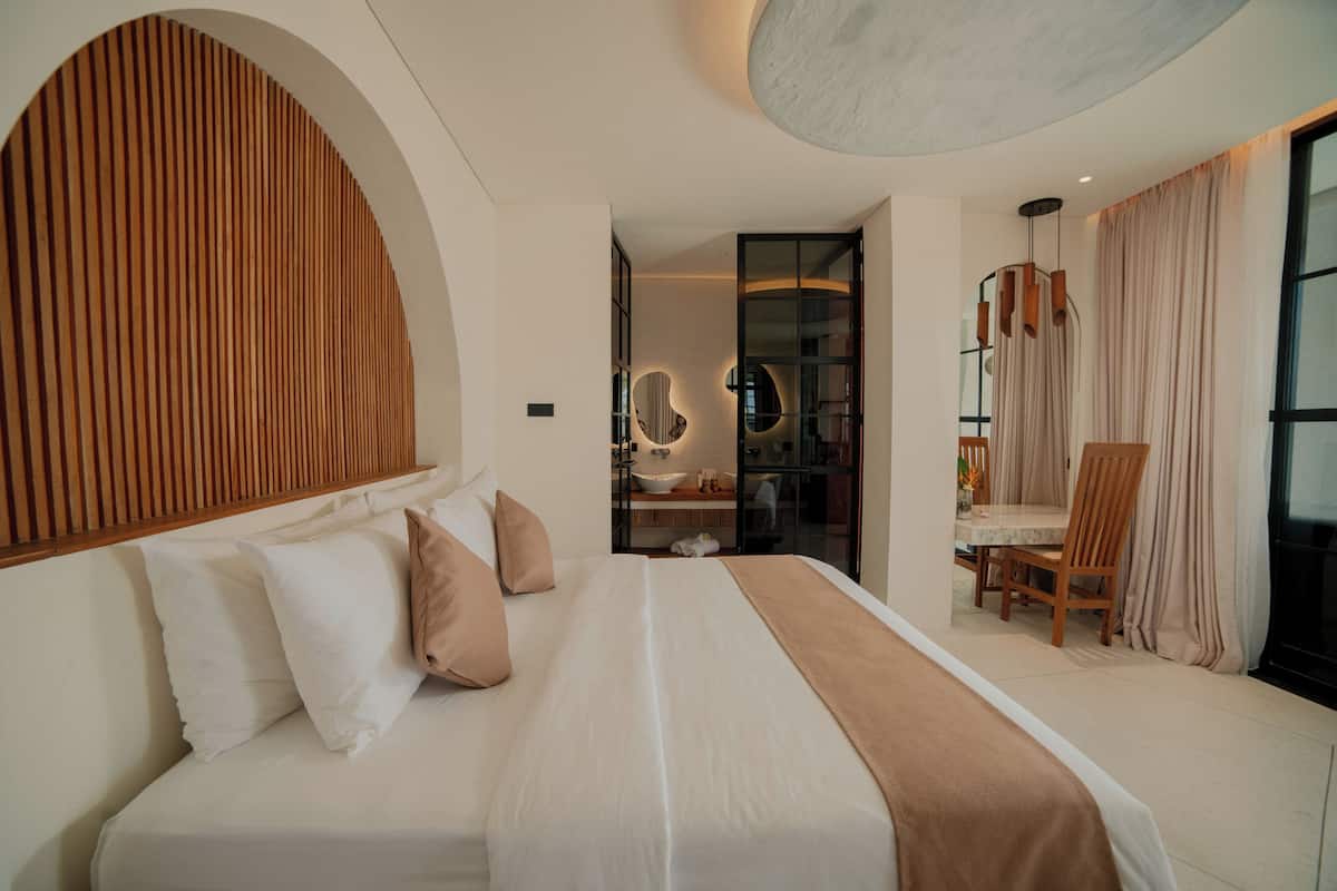 Ein modernes Hotelzimmer mit einem großen Bett, einem Holzkopfende, einer Sitzecke mit Tisch und Stühlen sowie einem Badezimmer mit Spiegel und Waschbecken.