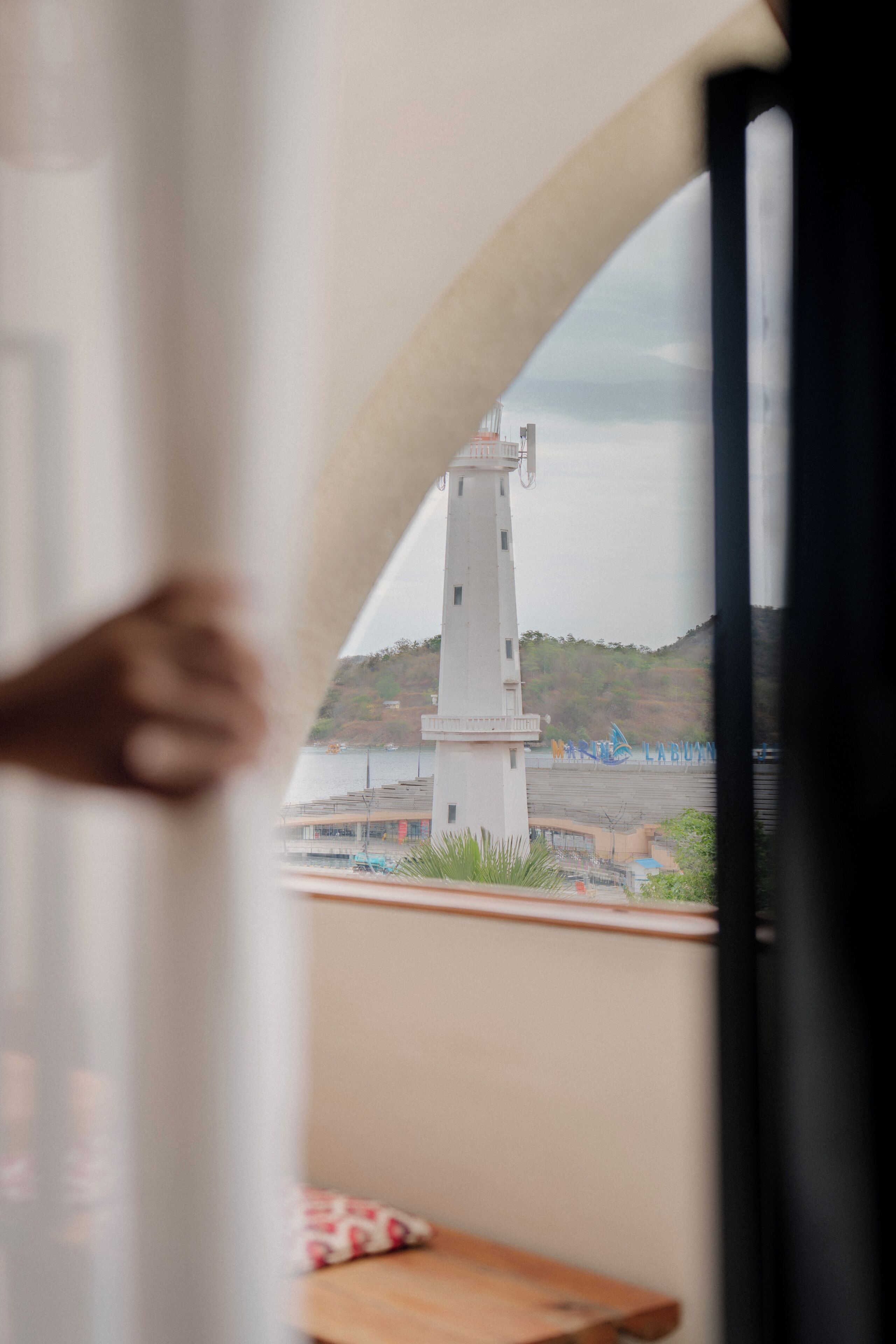 Vista de un faro a través de una ventana, una mesa de madera y una cortina blanca.