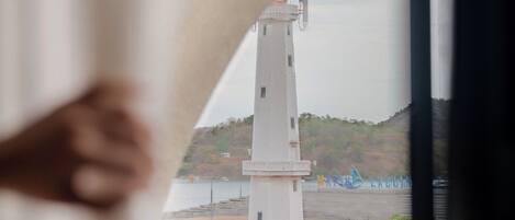 Vista de un faro a través de una ventana, una mesa de madera y una cortina blanca.