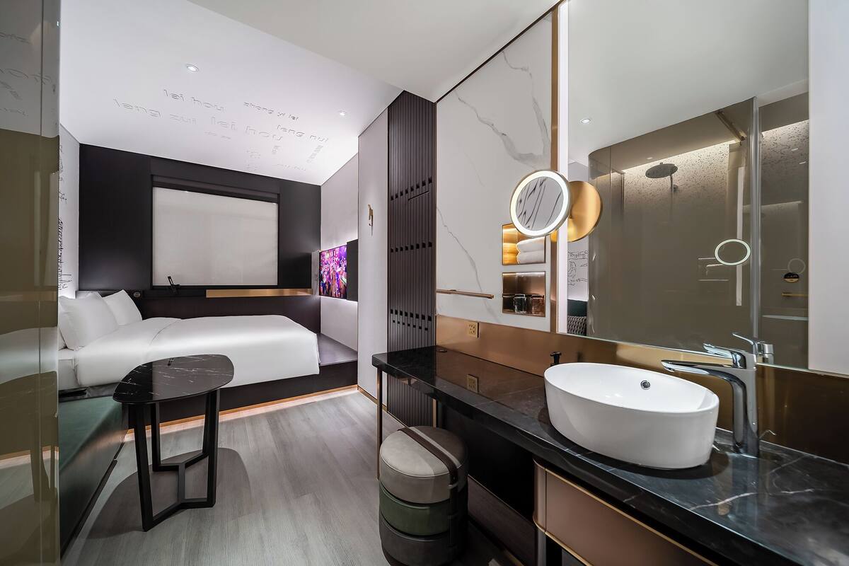 Ein modernes Hotelzimmer mit einem großen Bett, einem runden Spiegel mit Beleuchtung, einem weißen Waschbecken und einem kleinen Tisch.