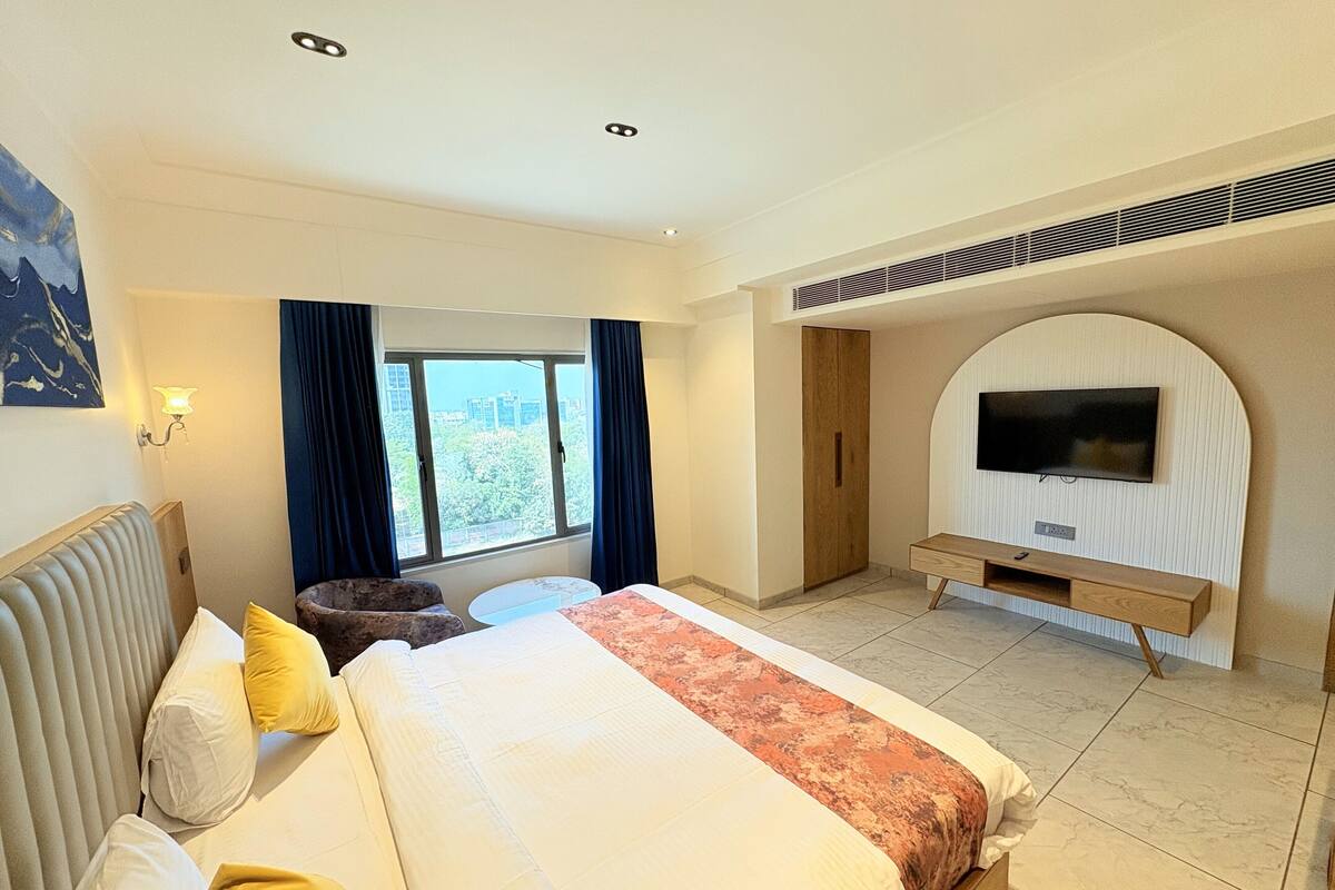 A hotel room with a large bed, a TV mounted on a curved wall, a window with curtains, and a view of buildings outside.