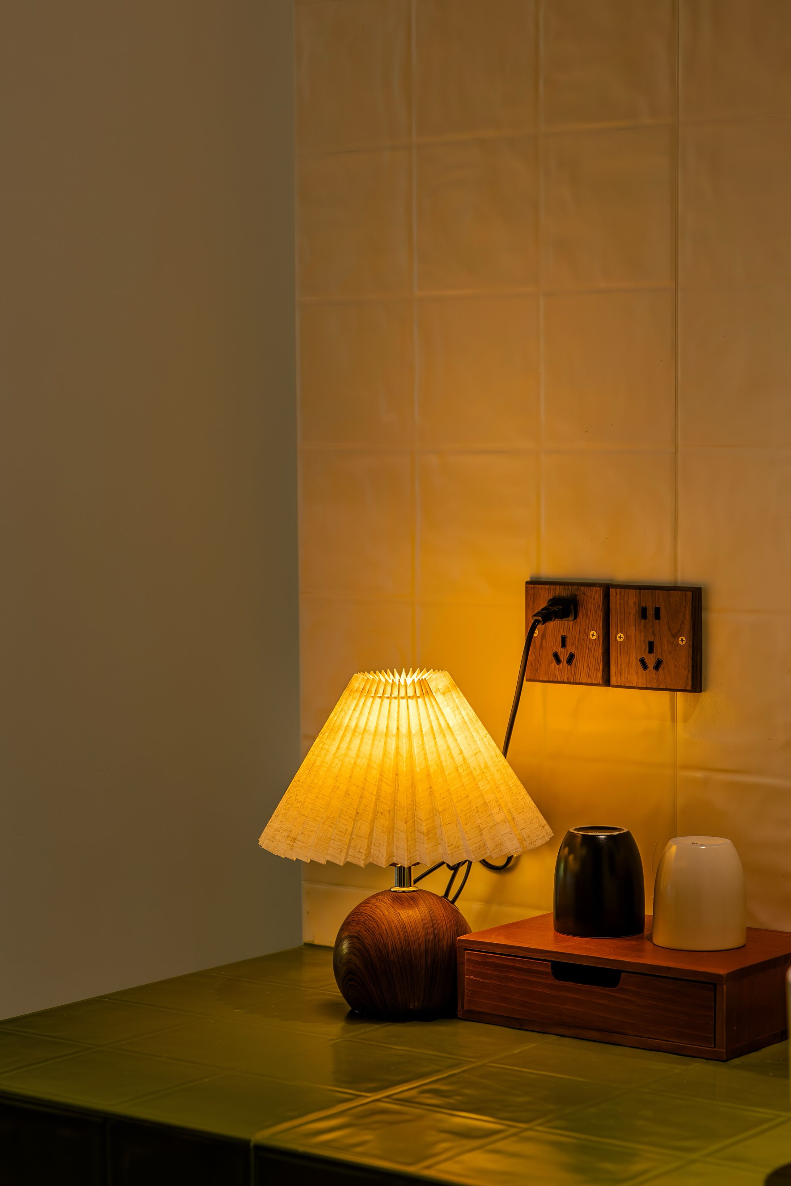 A table with a lamp, a vase, and a power outlet on a wall.