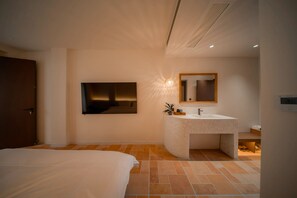 Une chambre moderne avec un grand lit, un téléviseur fixé au mur et une salle de bain équipée d’un meuble-lavabo et d’un évier.