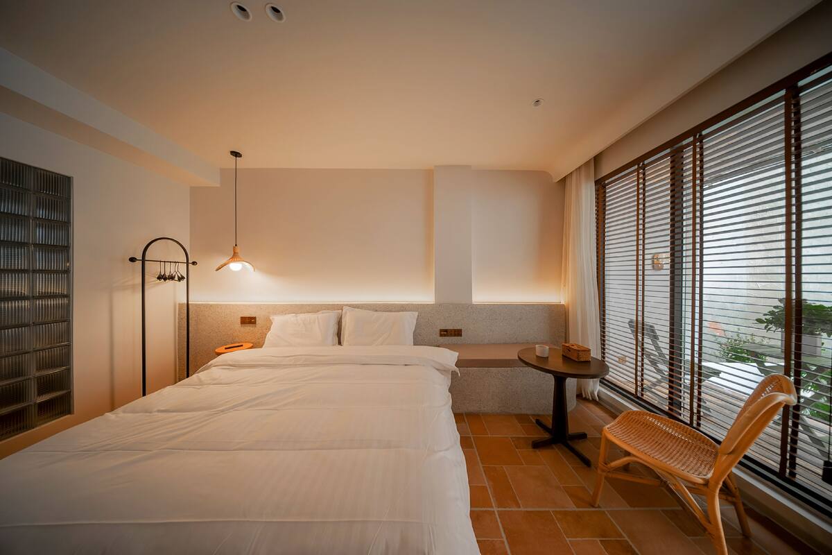 Quarto de hotel com uma cama grande, uma mesa pequena, uma cadeira e uma janela com persianas.