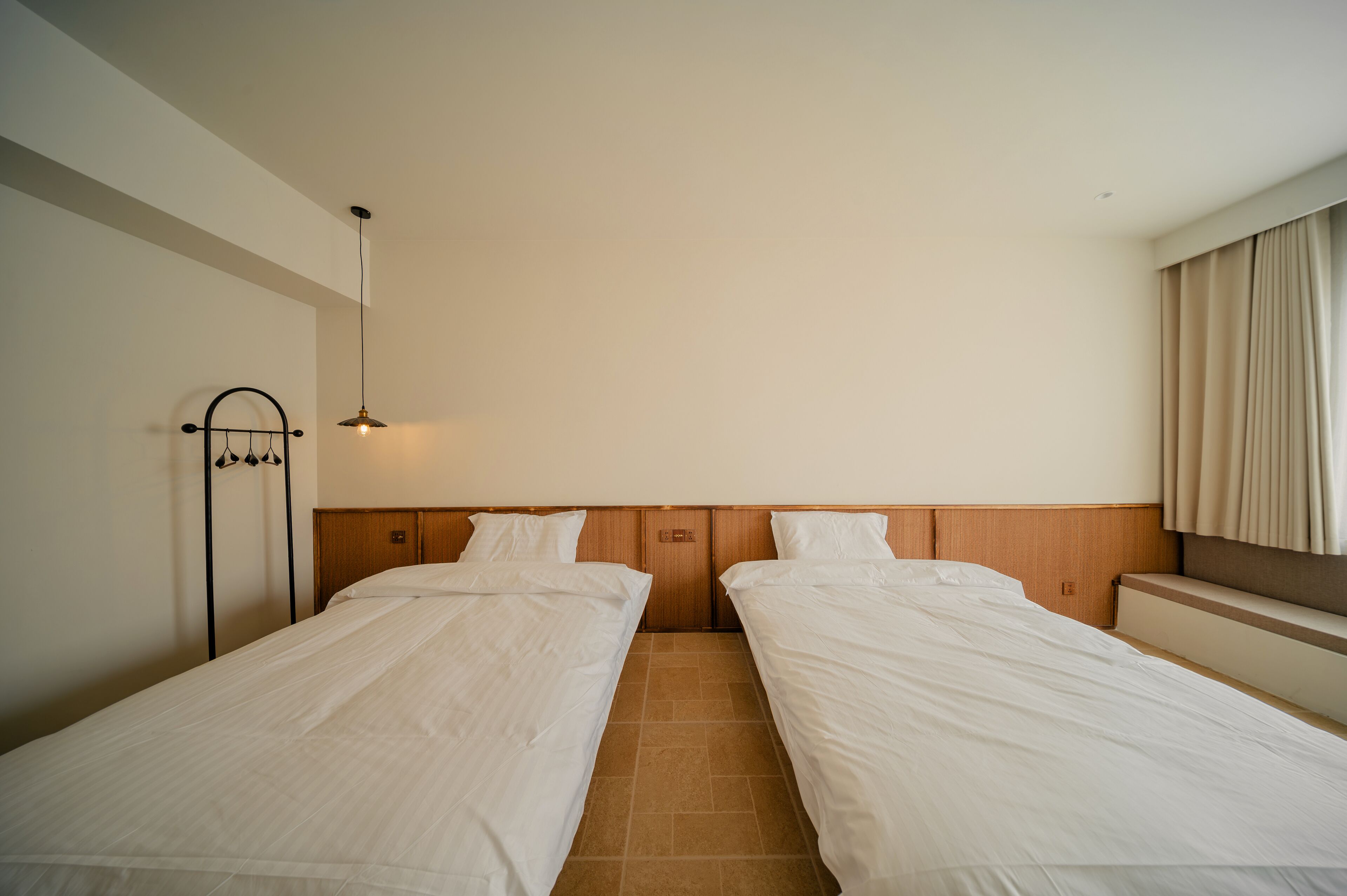 Una habitación de hotel con dos camas individuales, cabecero de madera, lámpara colgante y una ventana con cortinas.