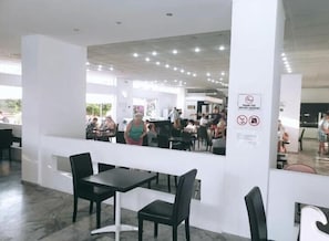 Un espacio público interior con mesas y sillas, un pilar blanco con un cartel, y una vista de personas al fondo.