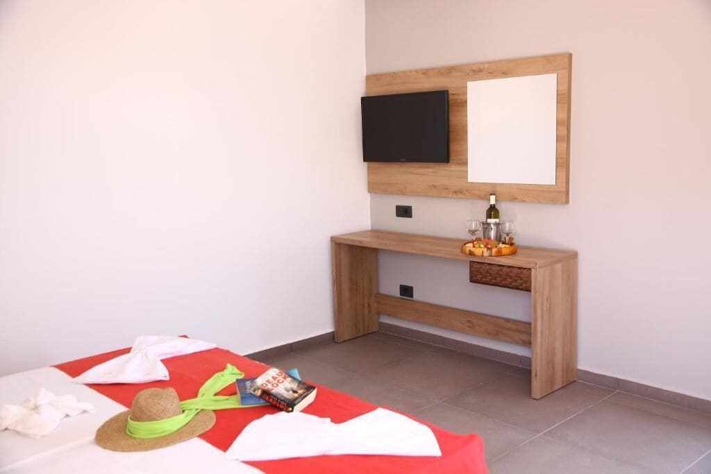 Un dormitorio con una cama, una mesita de noche con una botella, un televisor montado en la pared y un pequeño banco de madera.
