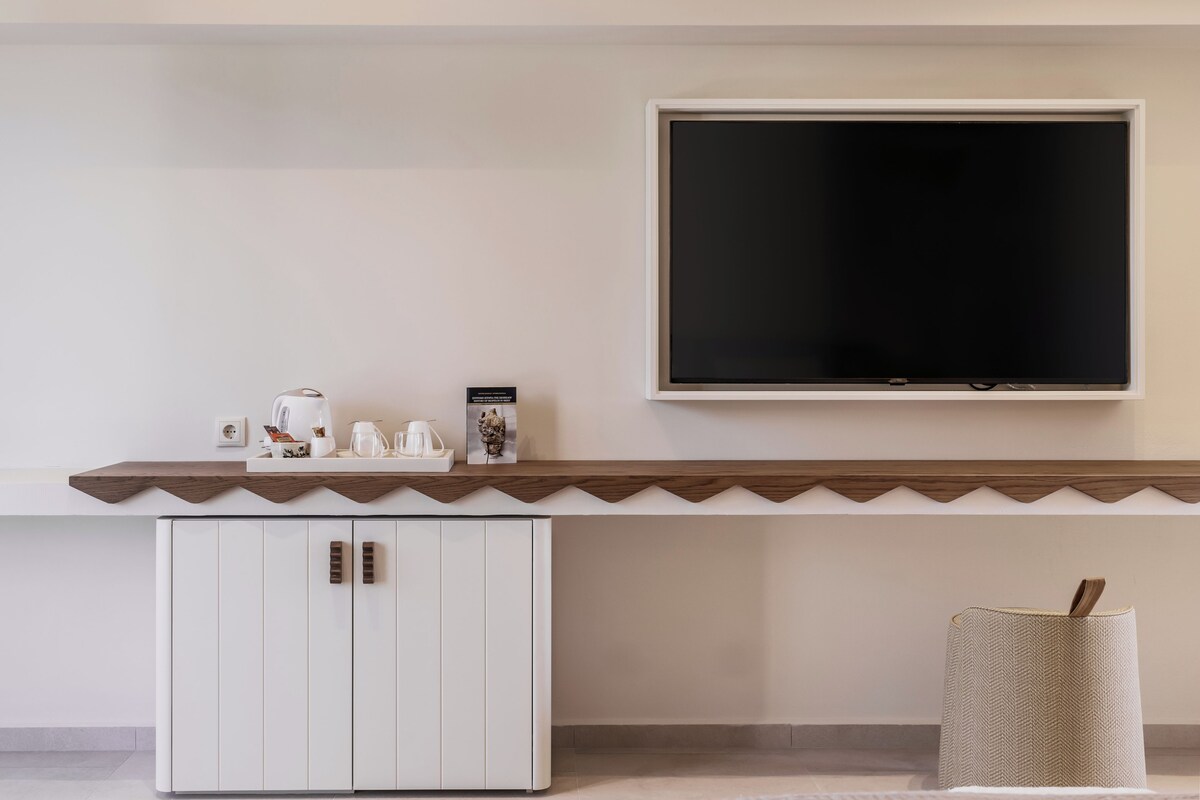 Ein modernes Zimmer mit einem wandmontierten Flachbildfernseher, einem Holzregal mit Kaffeemaschine und weiteren Gegenständen, einem weißen Schrank und einem gewebten Korb.