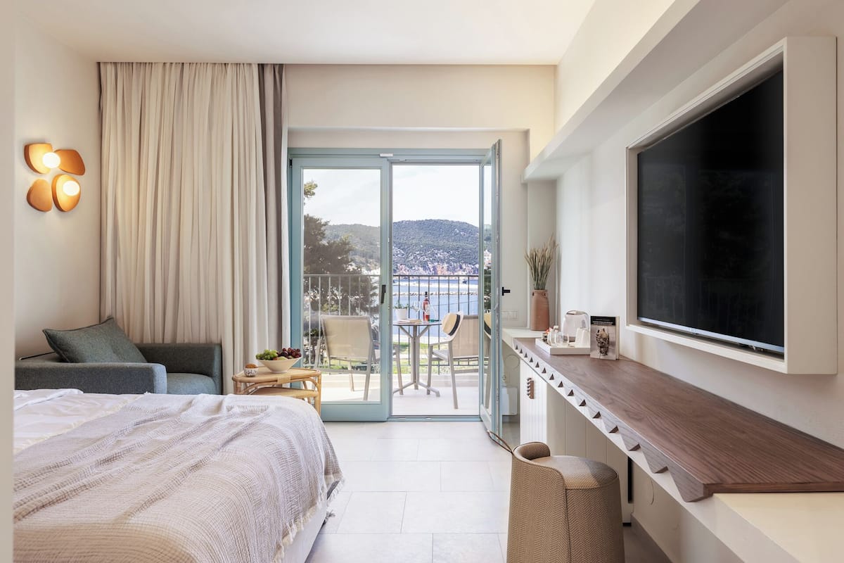 Una habitación de hotel con cama, escritorio con televisor de pantalla plana, balcón con vista al mar y zona de comedor con mesa y sillas.