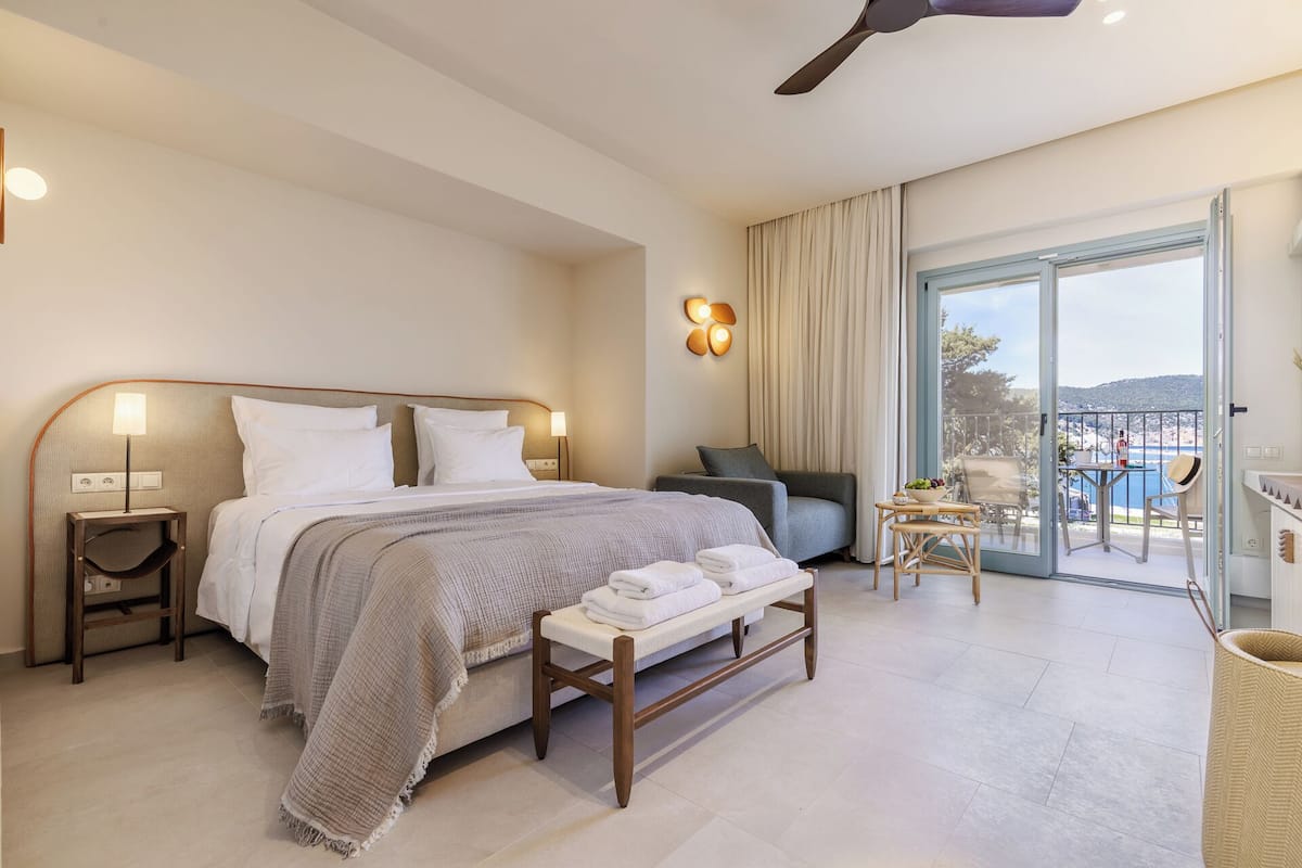 Habitación de hotel con cama, mesitas de noche, un banco, una silla, una pequeña mesa con un cuenco de fruta y vista al mar a través de un ventanal.
