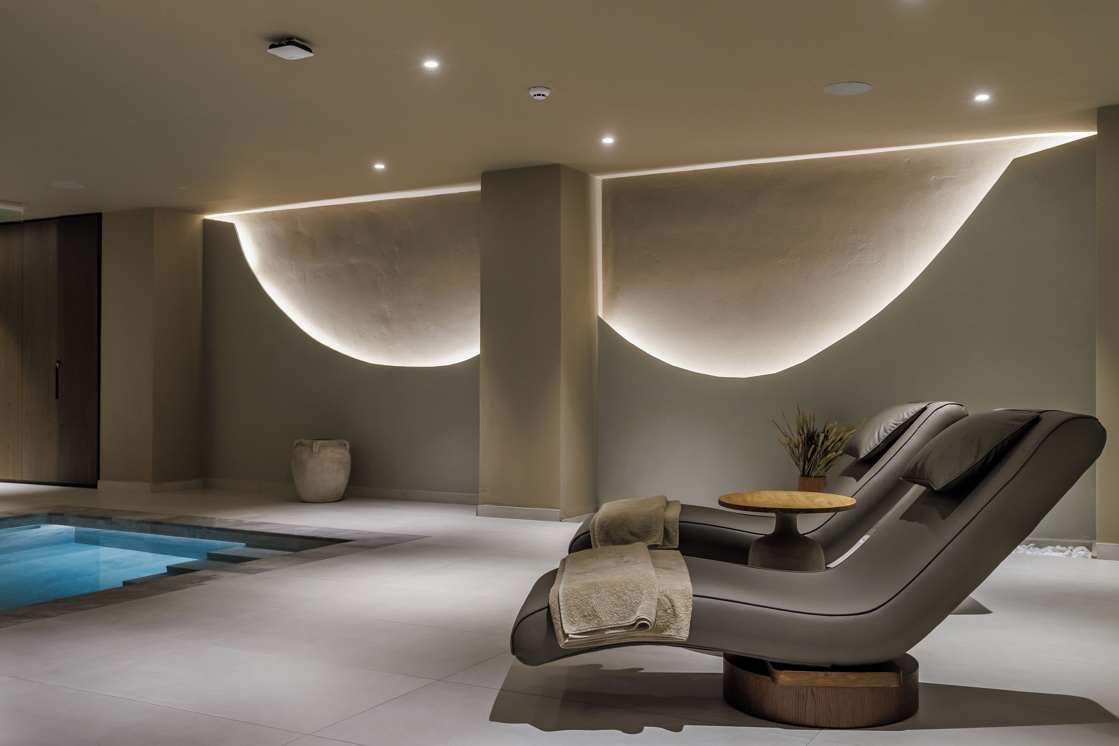 A modern spa room with a massage chair, a small table with a plant, and a swimming pool.