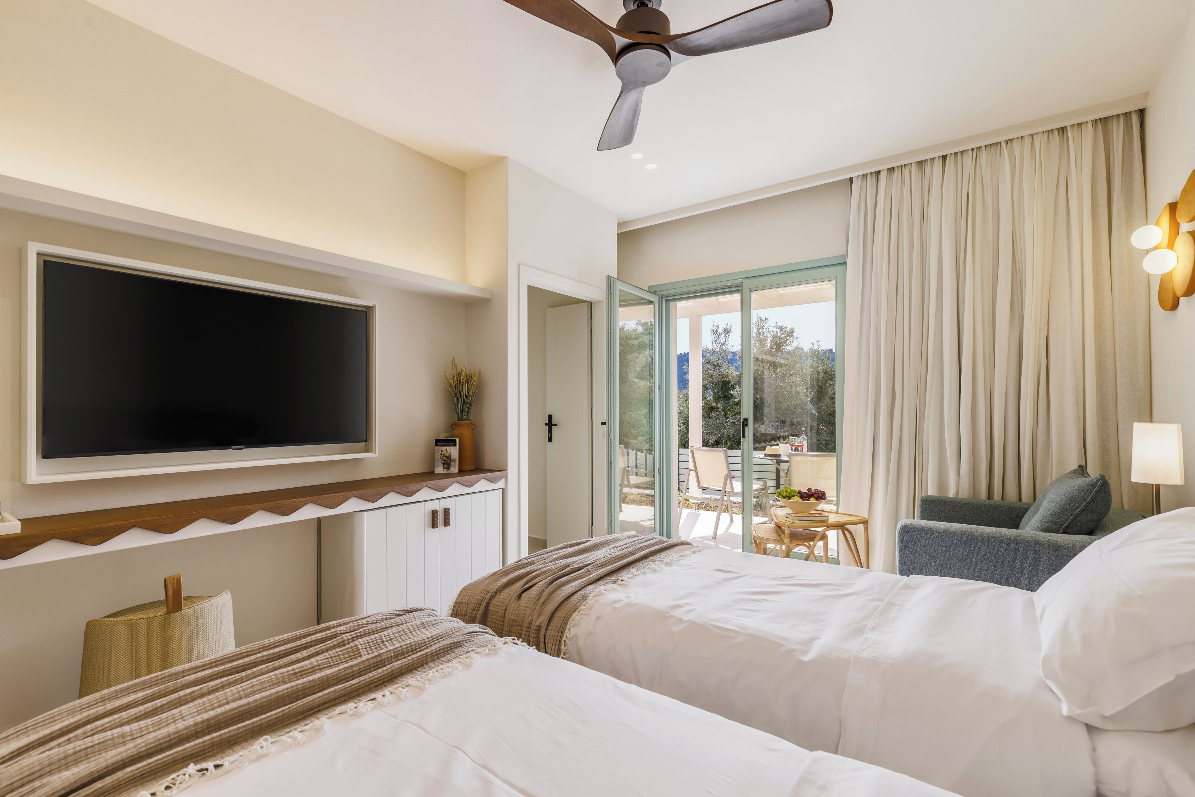 Ein Hotelzimmer mit Bett, Fernseher, Deckenventilator, einem Balkon mit Tisch und Stühlen sowie Blick ins Freie.