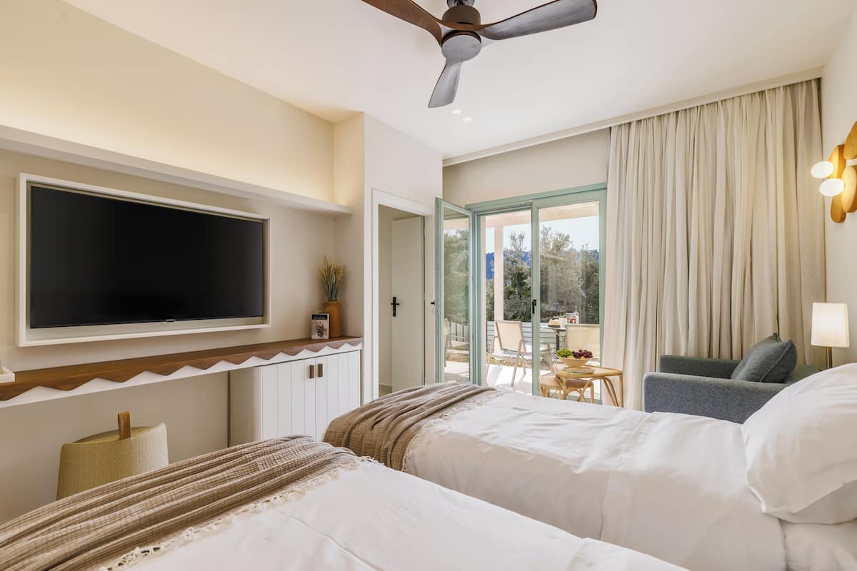 Habitación de hotel con cama, televisor, ventilador de techo, balcón con mesa y sillas, y vistas al exterior.