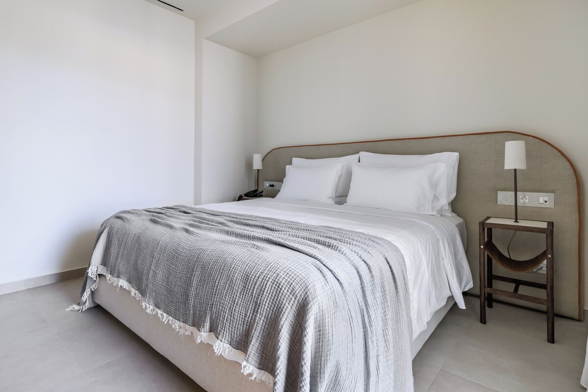 Un lit bien fait, recouvert d’une couverture grise, avec une lampe de chevet et une petite table de nuit en bois.