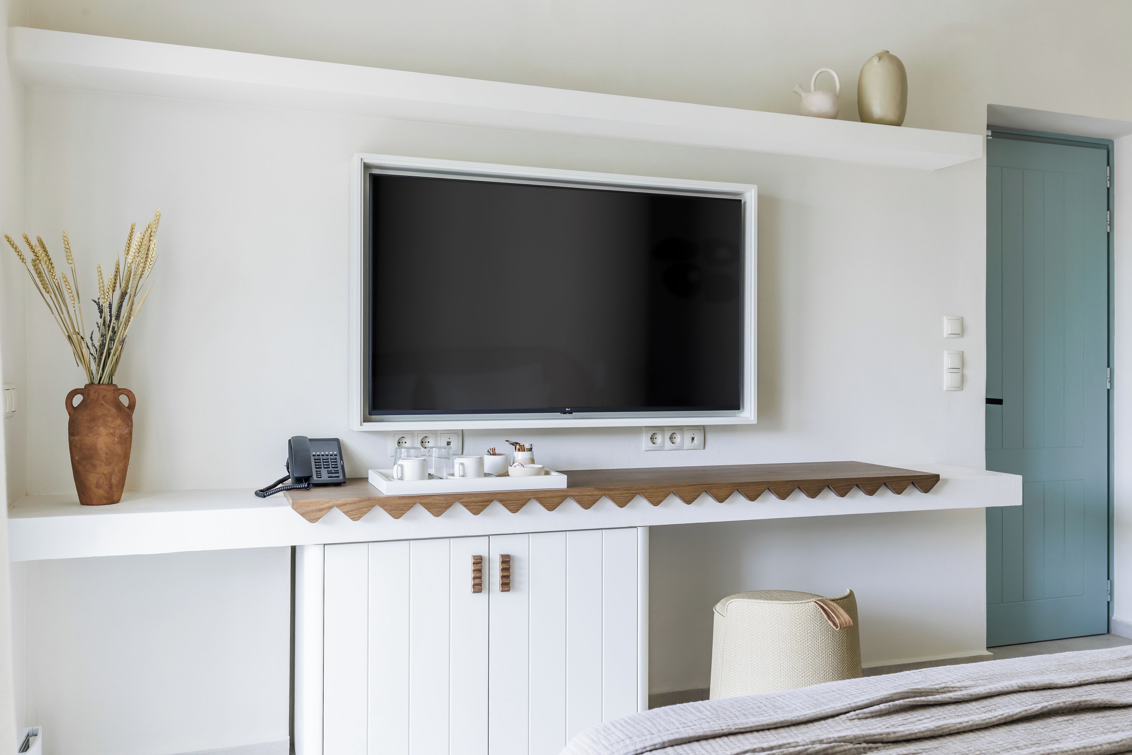 Ein modernes Hotelzimmer mit einem großen Flachbildfernseher an der Wand, einem kleinen Schreibtisch mit Telefon und Dekoration sowie einer Tür mit einem türkisfarbenen Einsatz.