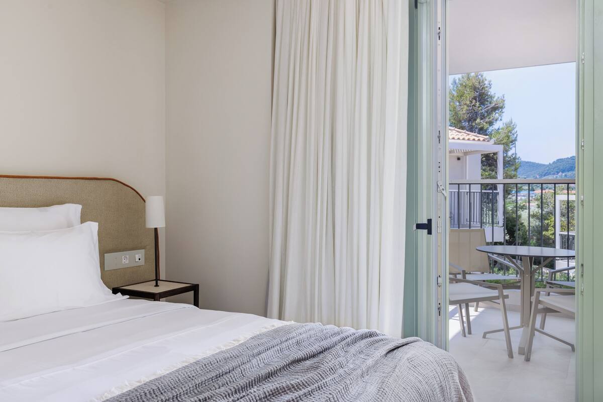 Habitación de hotel con cama, mesita de noche, lámpara y vistas a un balcón exterior con mesas y sillas.
