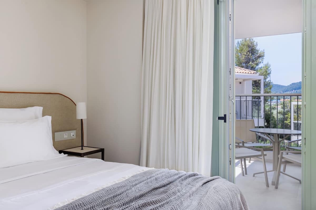 Ein Hotelzimmer mit Bett, Nachttisch, Lampe und Blick auf einen Außenbalkon mit Tischen und Stühlen.