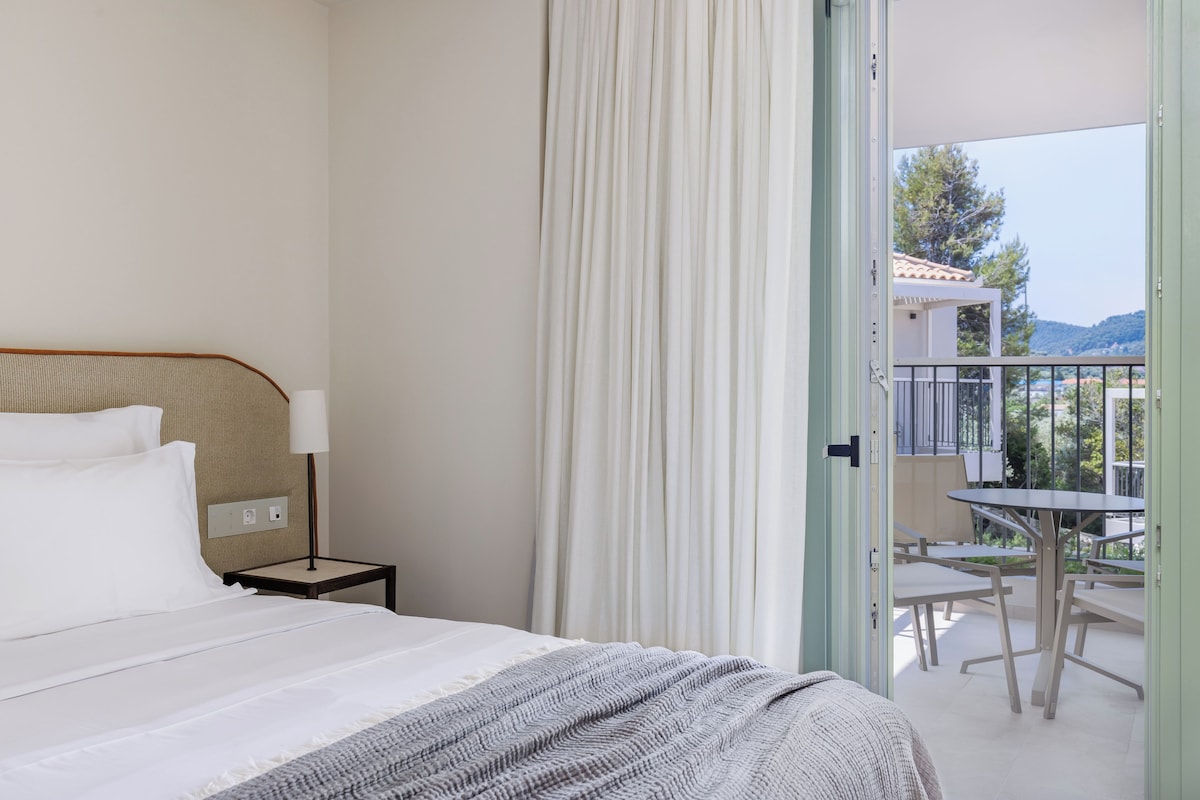 Ein Hotelzimmer mit Bett, Nachttisch, Lampe und Blick auf einen Außenbalkon mit Tischen und Stühlen.