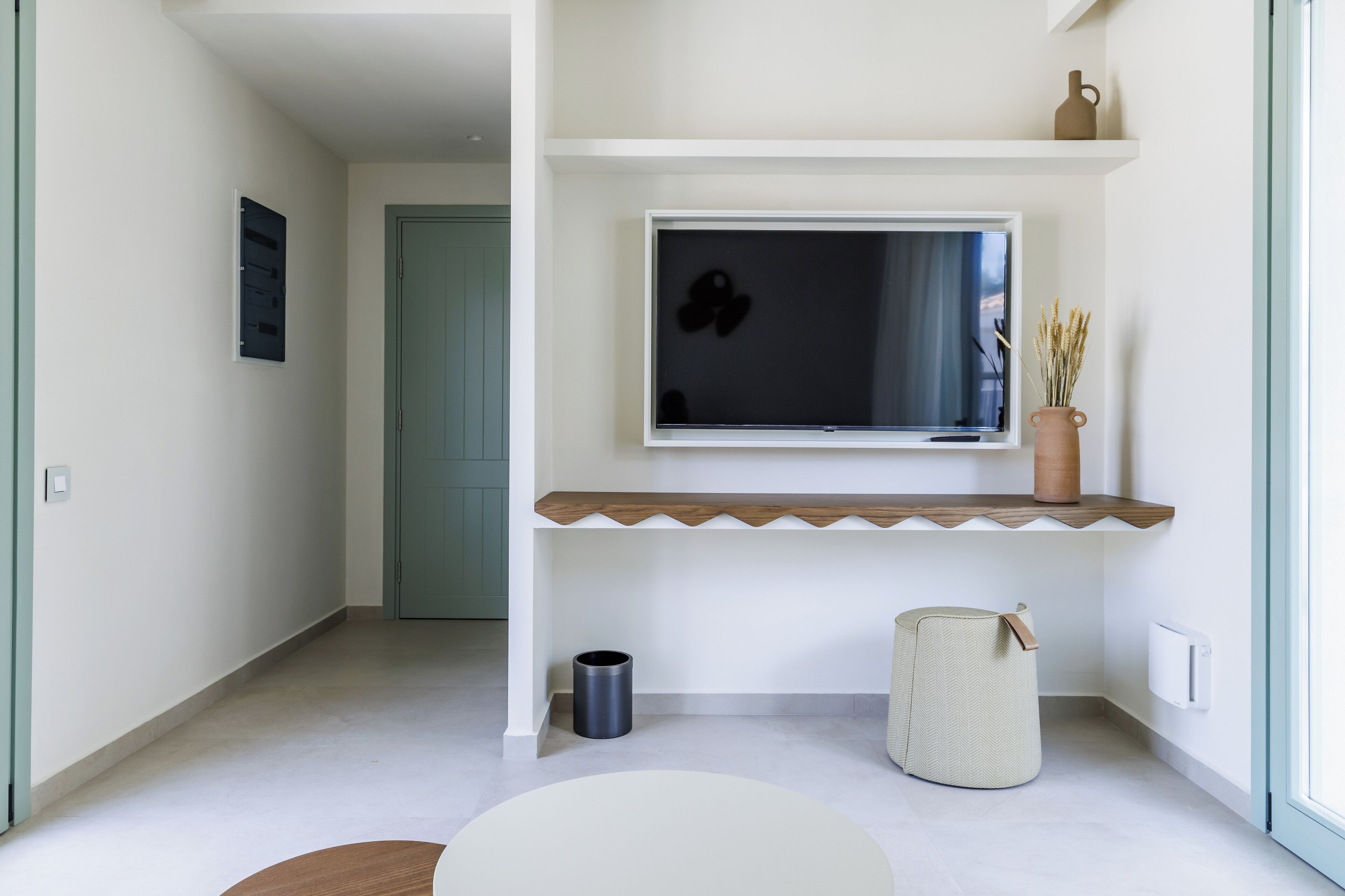A modern living room with a flat-screen TV on a wooden shelf, a small trash bin, a woven basket, and a door in the background.
