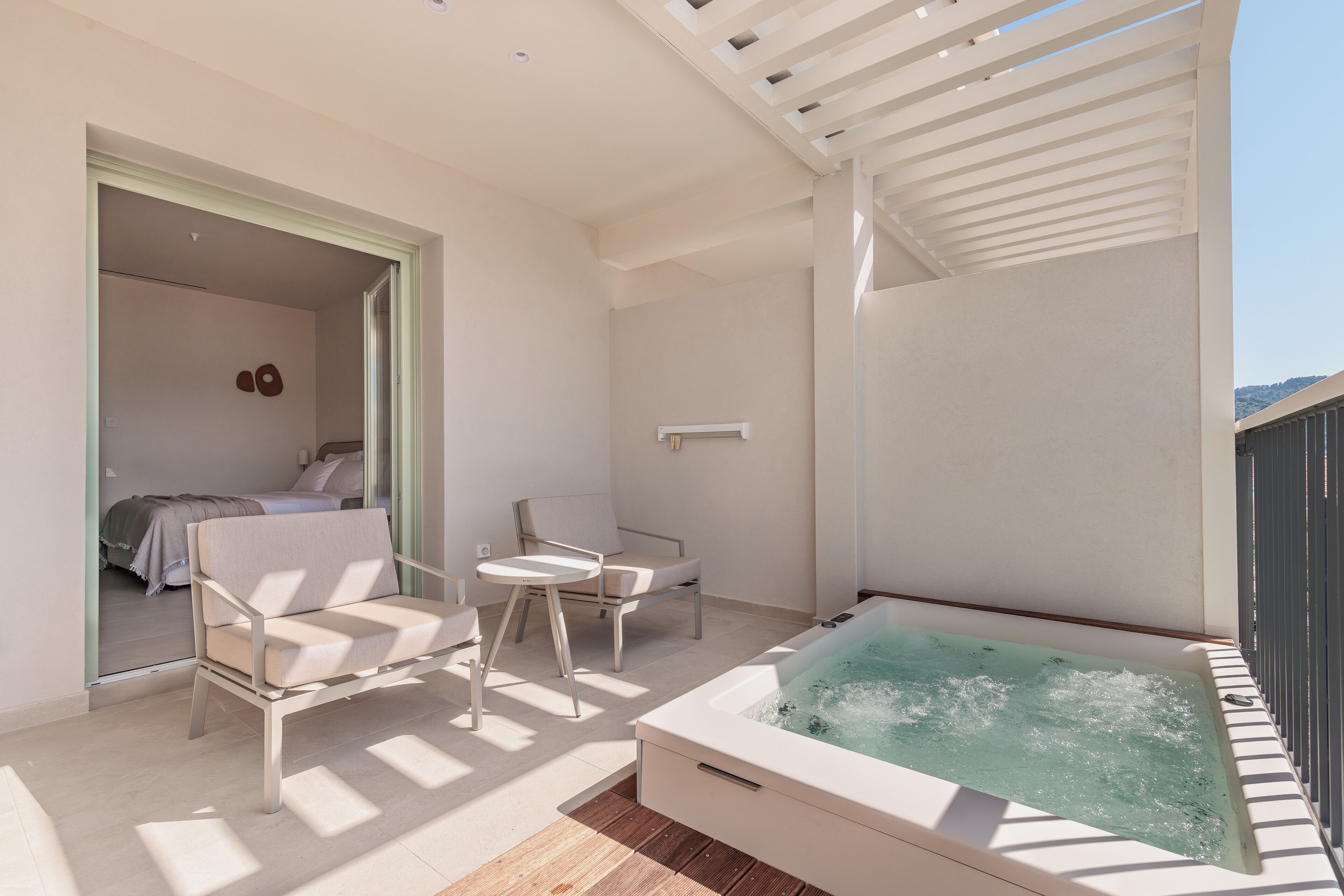 A balcony with a hot tub, two chairs, and a bed in the adjacent room.