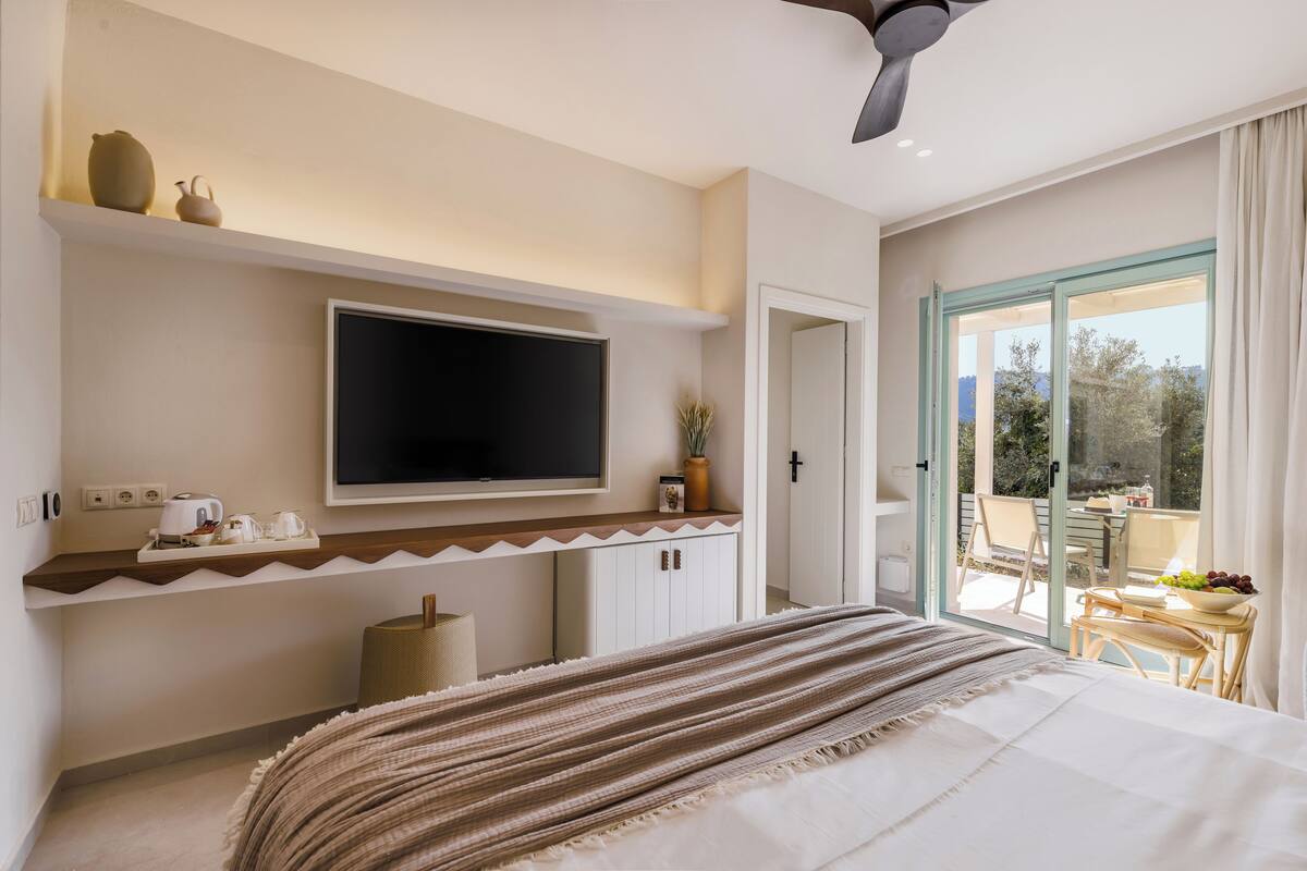 Un dormitorio con cama, televisor montado en la pared, una mesita con frutero y vistas al exterior a través de una puerta de cristal.