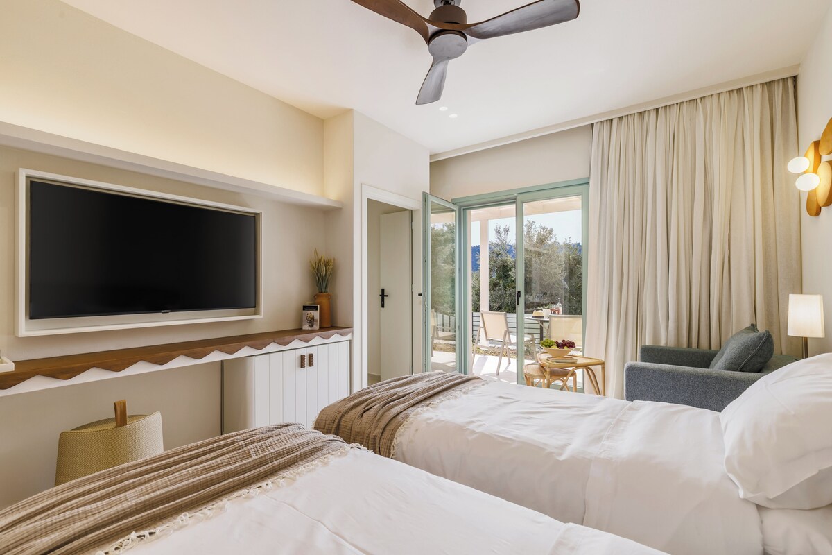 Une chambre d’hôtel équipée d’un lit, d’une télévision, d’un ventilateur de plafond, d’un balcon avec une table et des chaises, et offrant une vue sur l’extérieur.