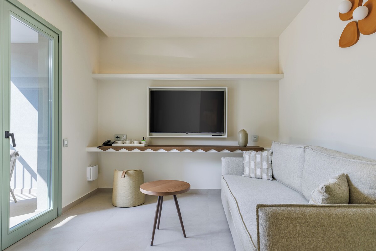 Ein modernes Wohnzimmer mit einer Couch, einem runden Holztisch, einem an der Wand befestigten Fernseher und einem großen Fenster.