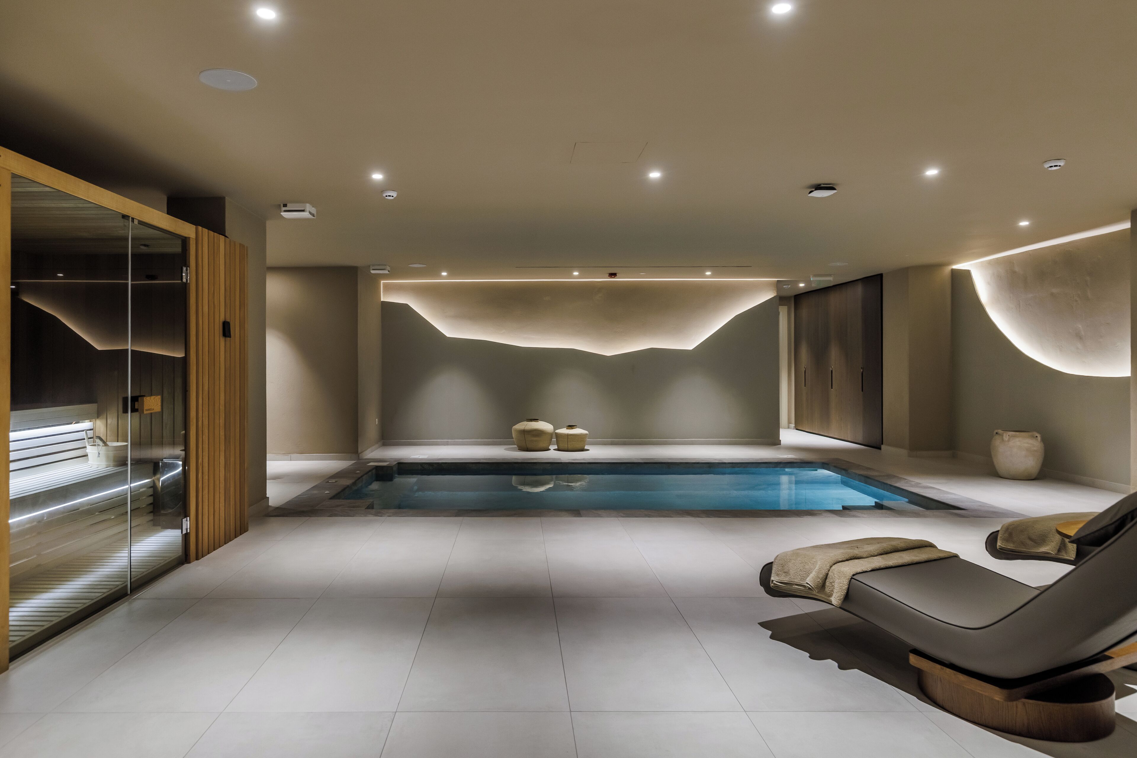 Une salle de spa moderne dotée d’une grande piscine, de deux chaises longues et d’une paroi en verre.