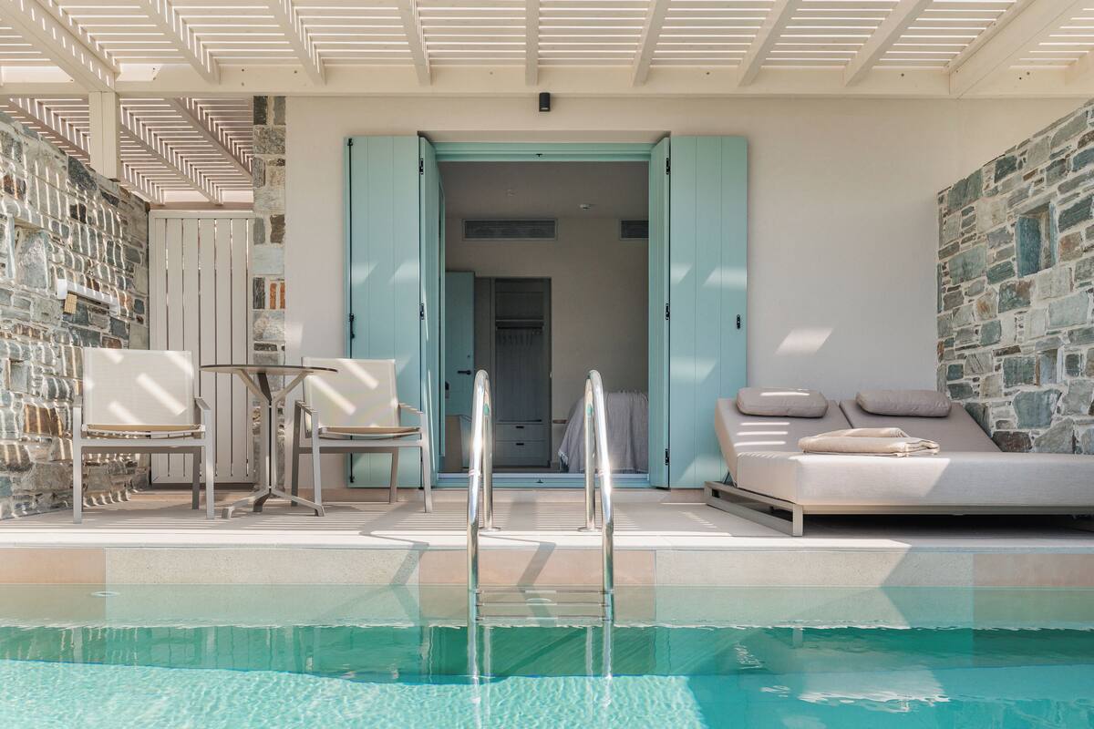 Zona de piscina con una silla de descanso, una mesa y una escalera que lleva a un espacio interior.