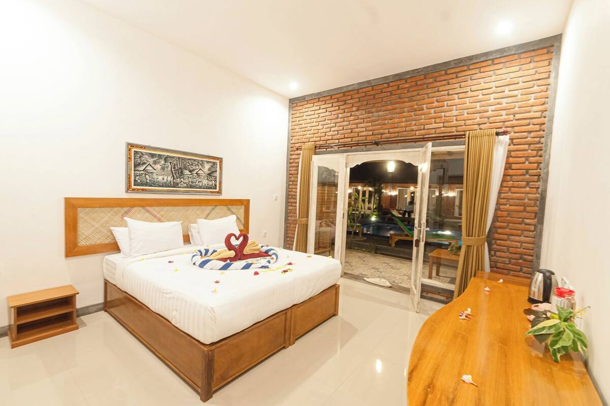 A hotel room with a bed, a wooden nightstand, a framed picture, and a view of an outdoor pool area.