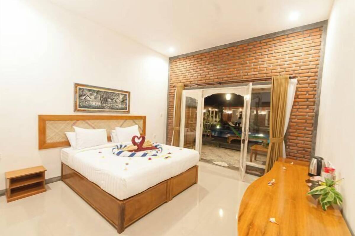 Un dormitorio con cama, mesita de noche de madera, pared decorativa de ladrillo, puerta de cristal que da a un balcón y una mesa de madera con una planta.