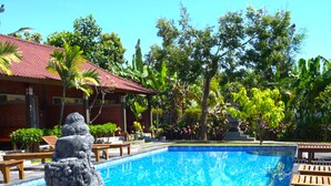 A swimming pool with a statue, surrounded by greenery and a building with a red roof. - Rumah Gajah by Kubukubali (Pemuteran)