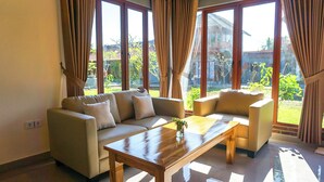A living room with a sofa, a coffee table, and a chair. There are large windows with curtains. - Rumah Gajah by Kubukubali (Pemuteran)